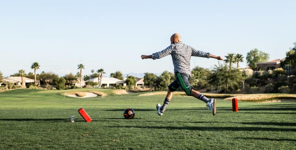 Joueur de footgolf en action, pied levé pour frapper la balle de football vers un trou sur un parcours de golf