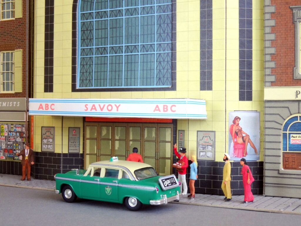 Diorama urbain recréant une rue animée avec un véhicule et des piétons devant un cinéma